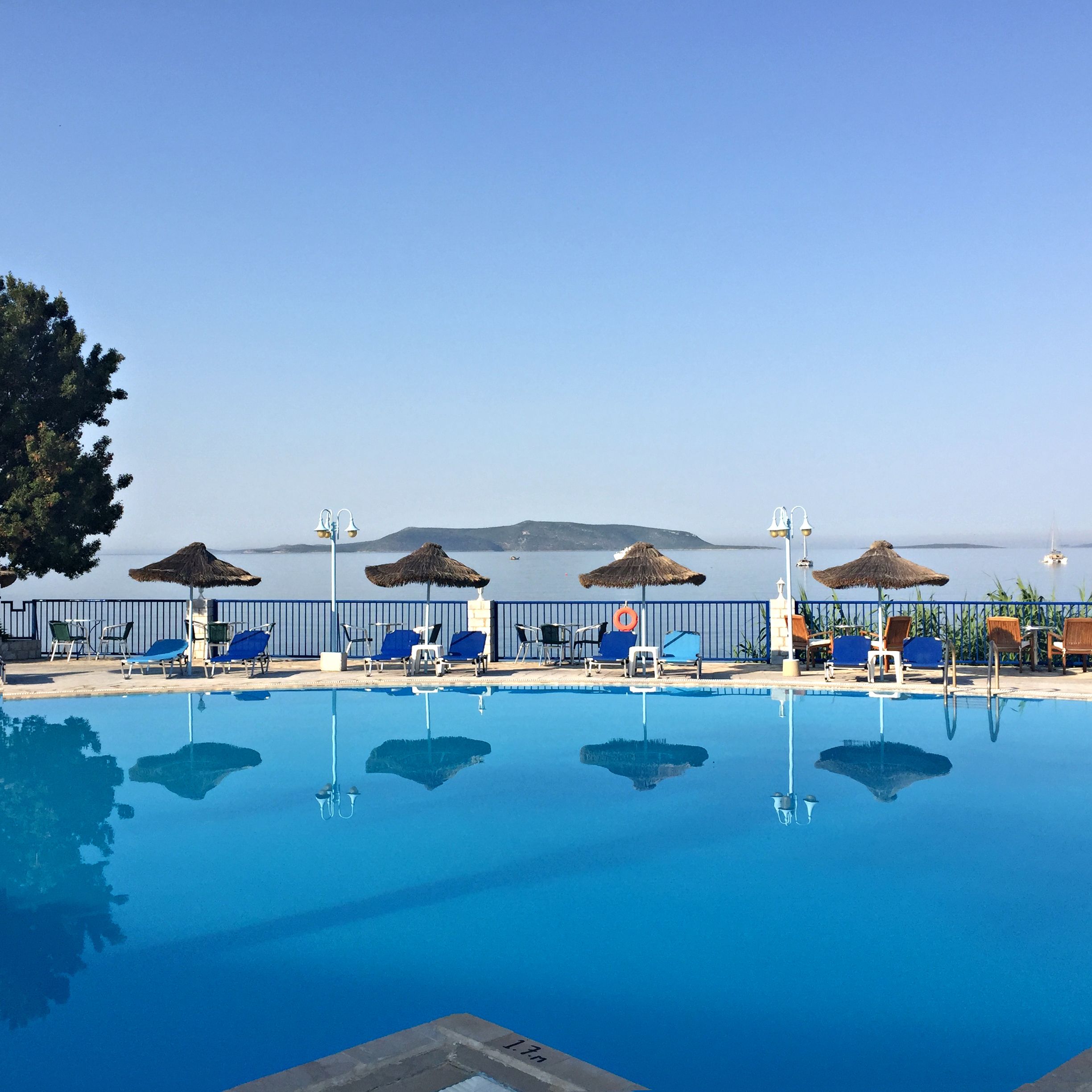 Una piscina con tumbonas y sombrillas, rodeada por una valla, con vista al mar y colinas distantes.