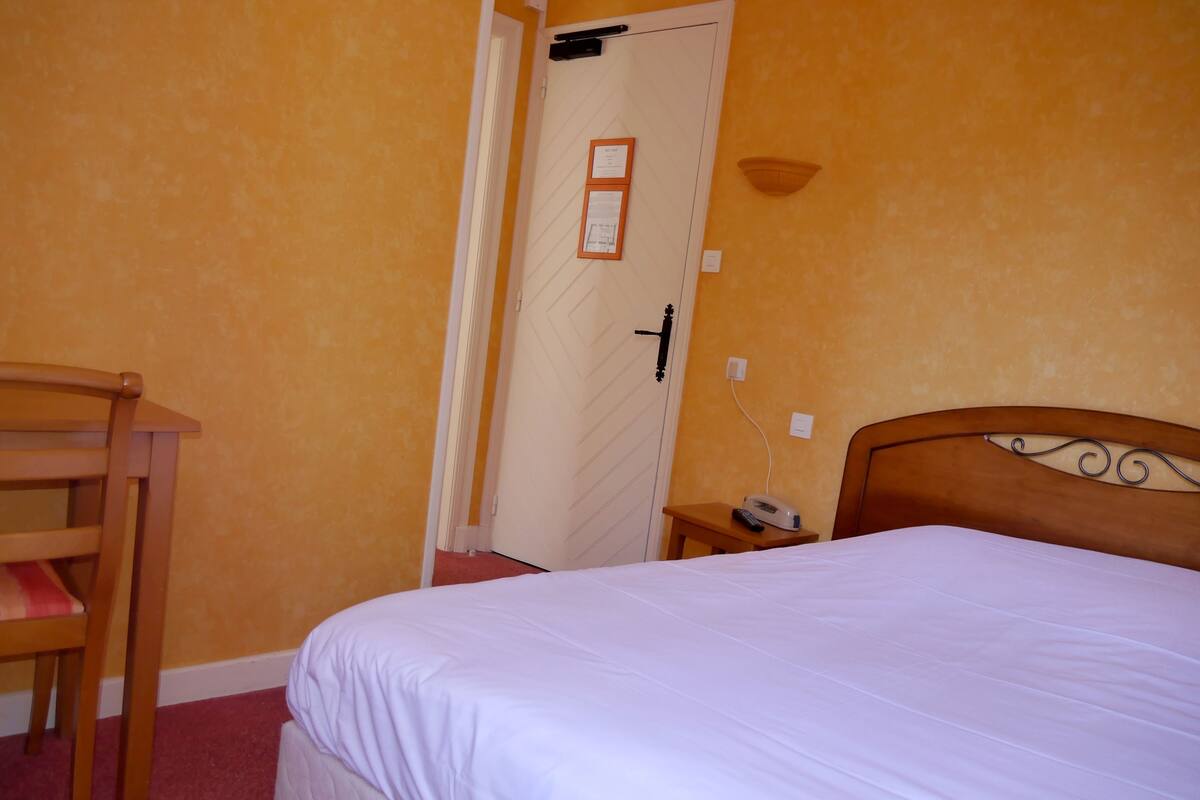 Une chambre d’hôtel avec un lit, une chaise en bois, une table de chevet avec un téléphone et une porte dotée d’un interrupteur pour la lumière.