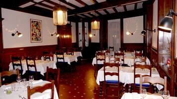Un restaurante con varias mesas y sillas, vigas de madera en el techo y luces fijadas a la pared.