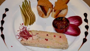 Een bord met diverse soorten eten, waaronder een stuk vis, brood, augurken en een klein schaaltje saus.