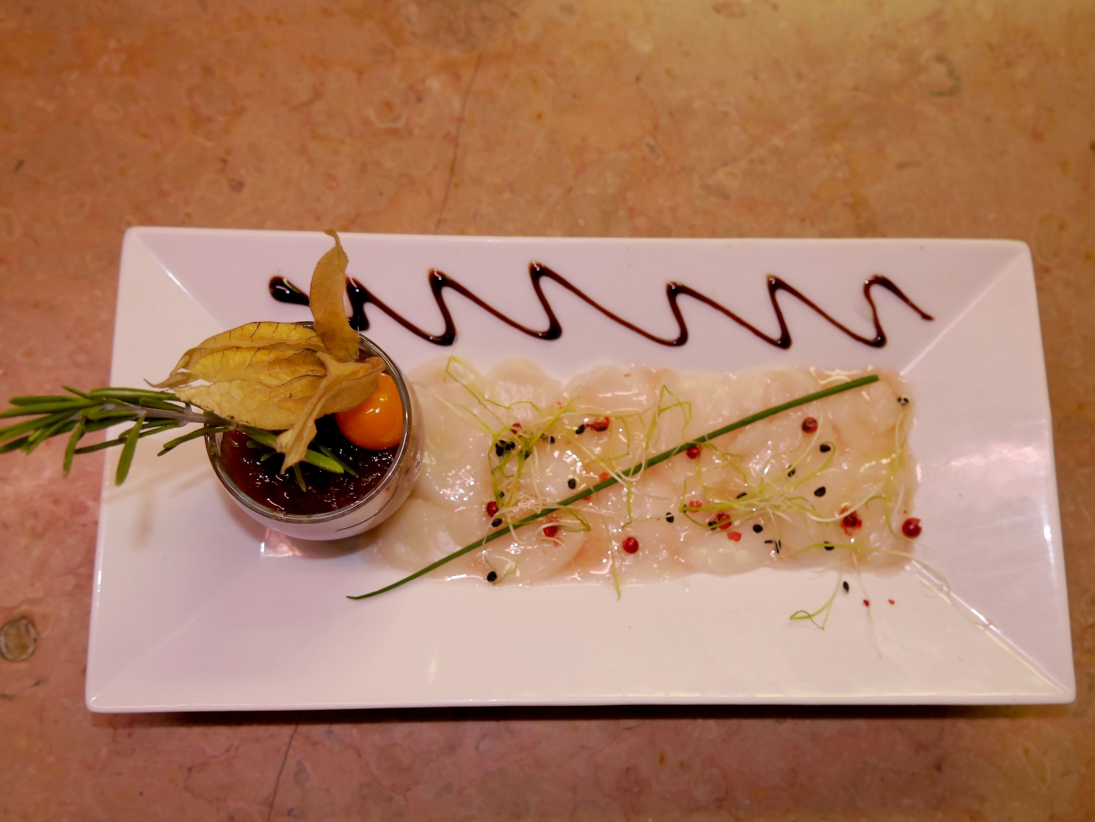 Une assiette rectangulaire blanche sur laquelle est disposé un plat de sashimi, garni d’oignons verts et de flocons de piment rouge, ainsi qu’un petit bol de sauce décoré d’une feuille.