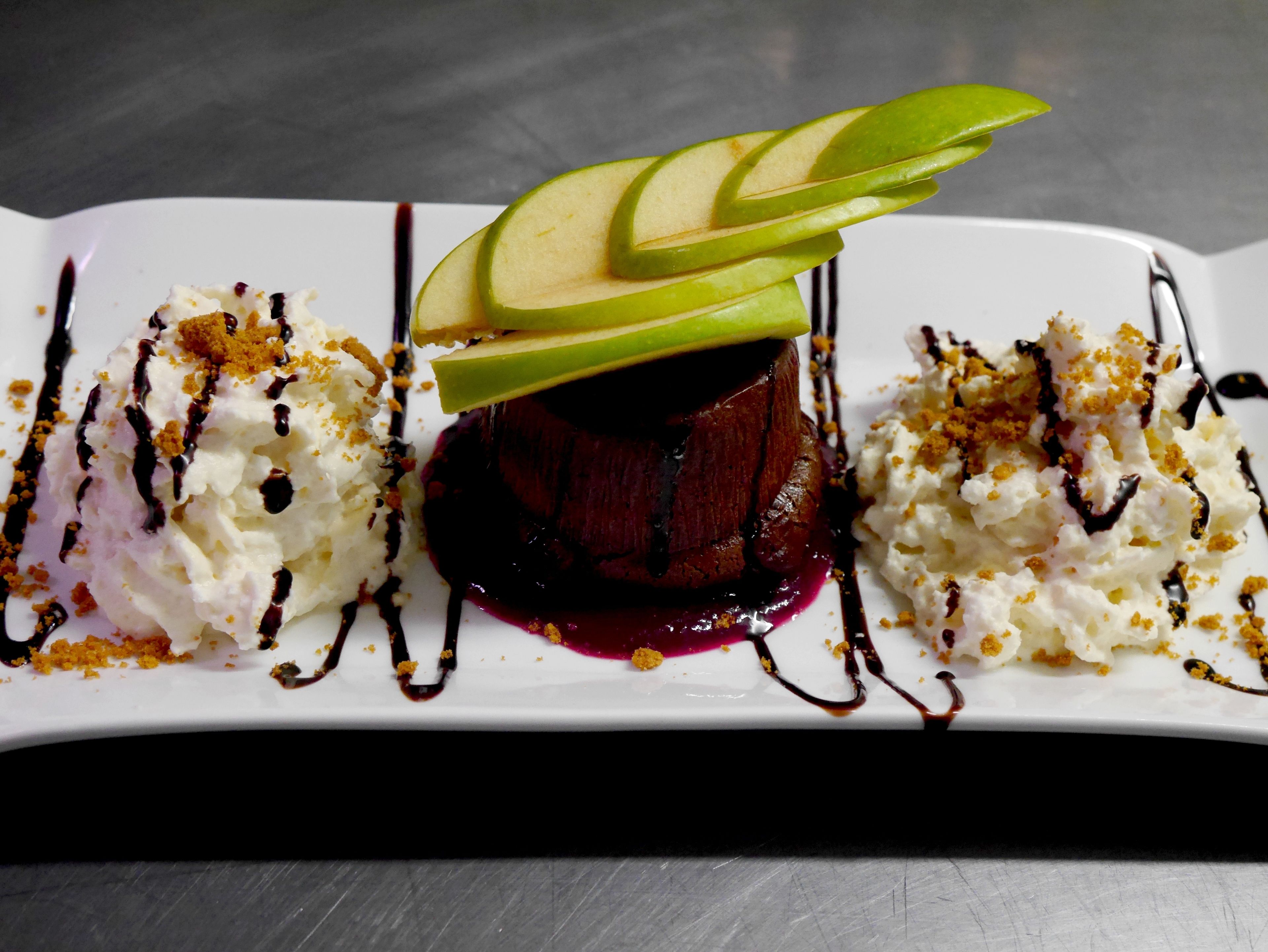 Um prato de sobremesa com um bolo vermelho escuro, fatias de maçã, chantilly e calda de chocolate.
