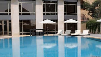 A swimming pool with white lounge chairs and umbrellas, surrounded by a building with large windows and a stone facade.