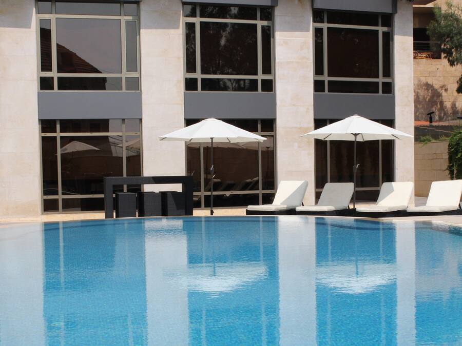 A swimming pool with white lounge chairs and umbrellas, surrounded by a building with large windows and a stone facade.