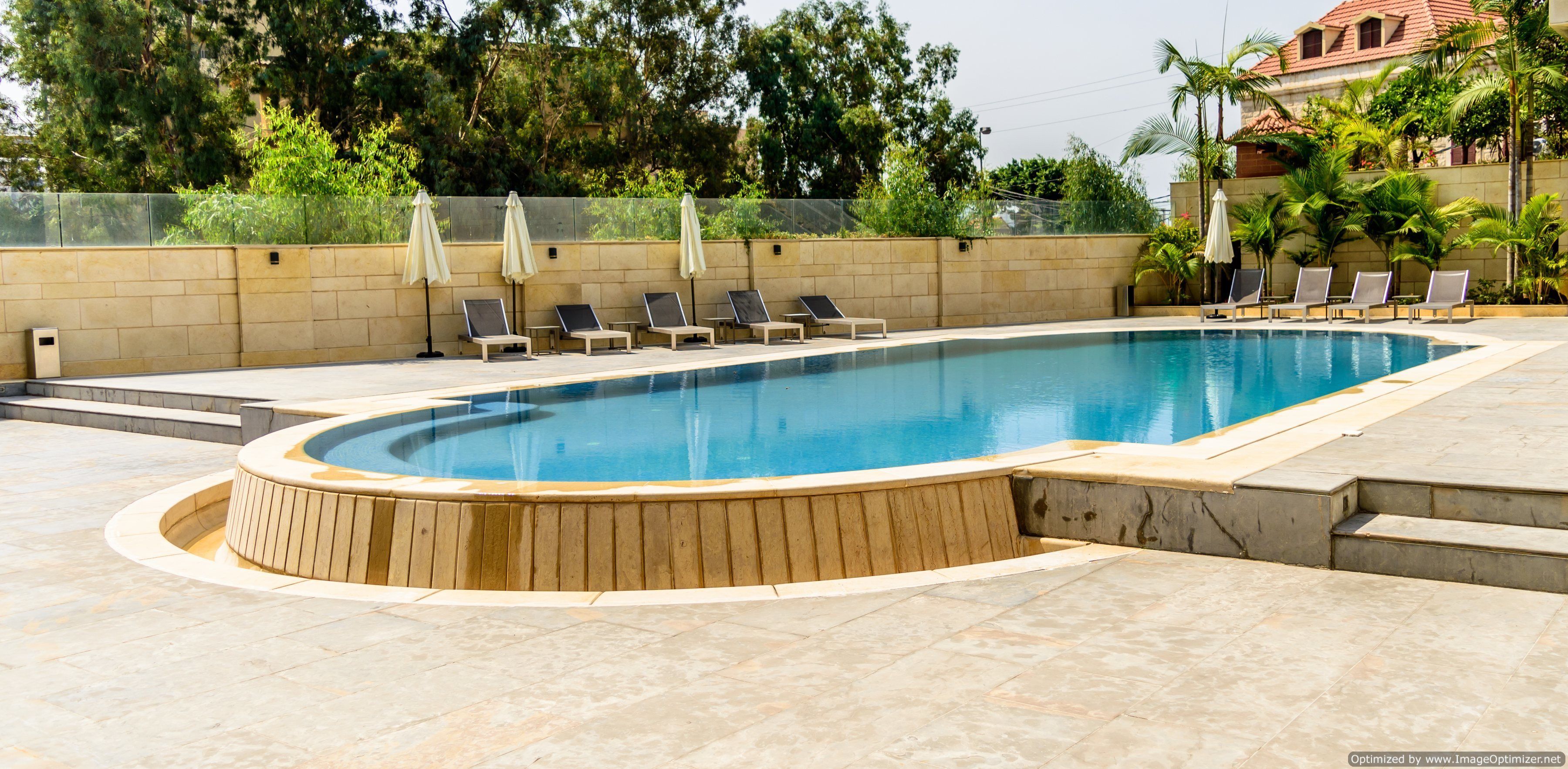 A swimming pool with lounge chairs and umbrellas, surrounded by a stone wall and greenery.