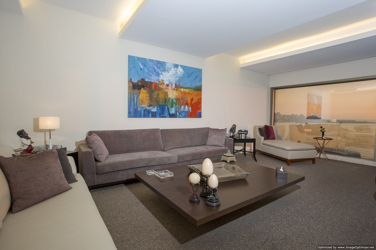 A modern living room with a gray sofa, a coffee table with decorative items, a painting on the wall, and a view through a large window.