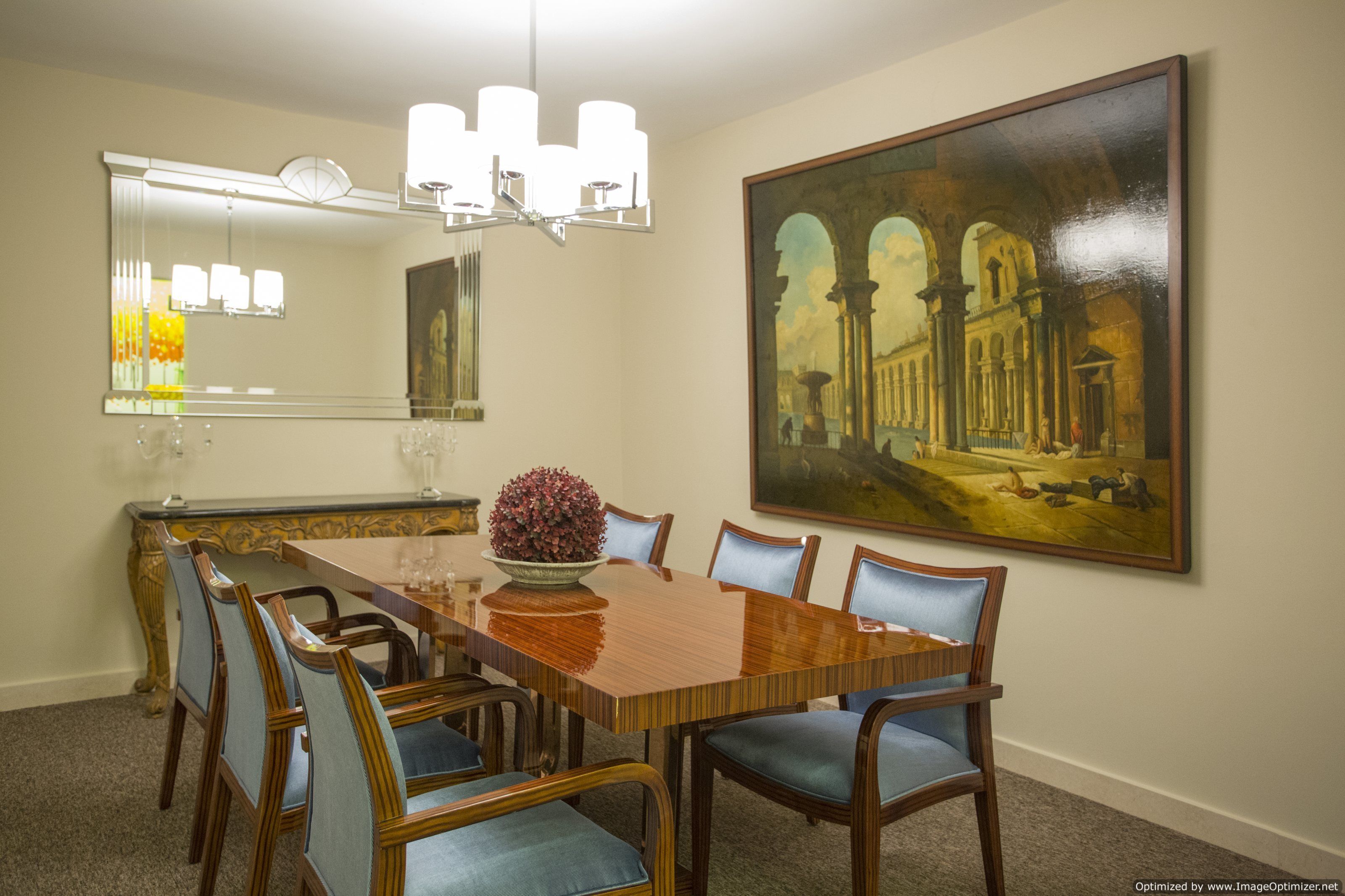 Comedor con una gran mesa de madera, seis sillas, una araña, un espejo grande y un cuadro de una escena arquitectónica.