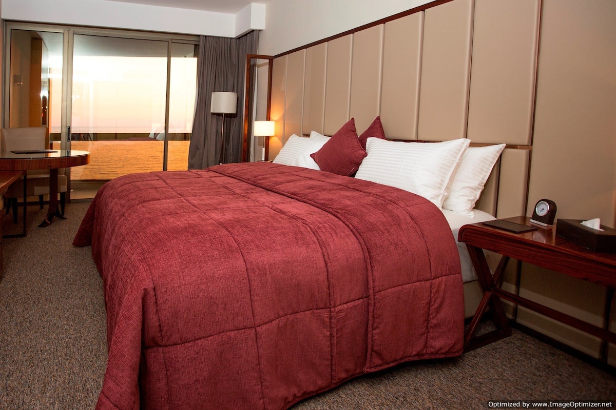 A hotel room with a large bed, a desk, a chair, a TV, and a sliding door to a balcony.