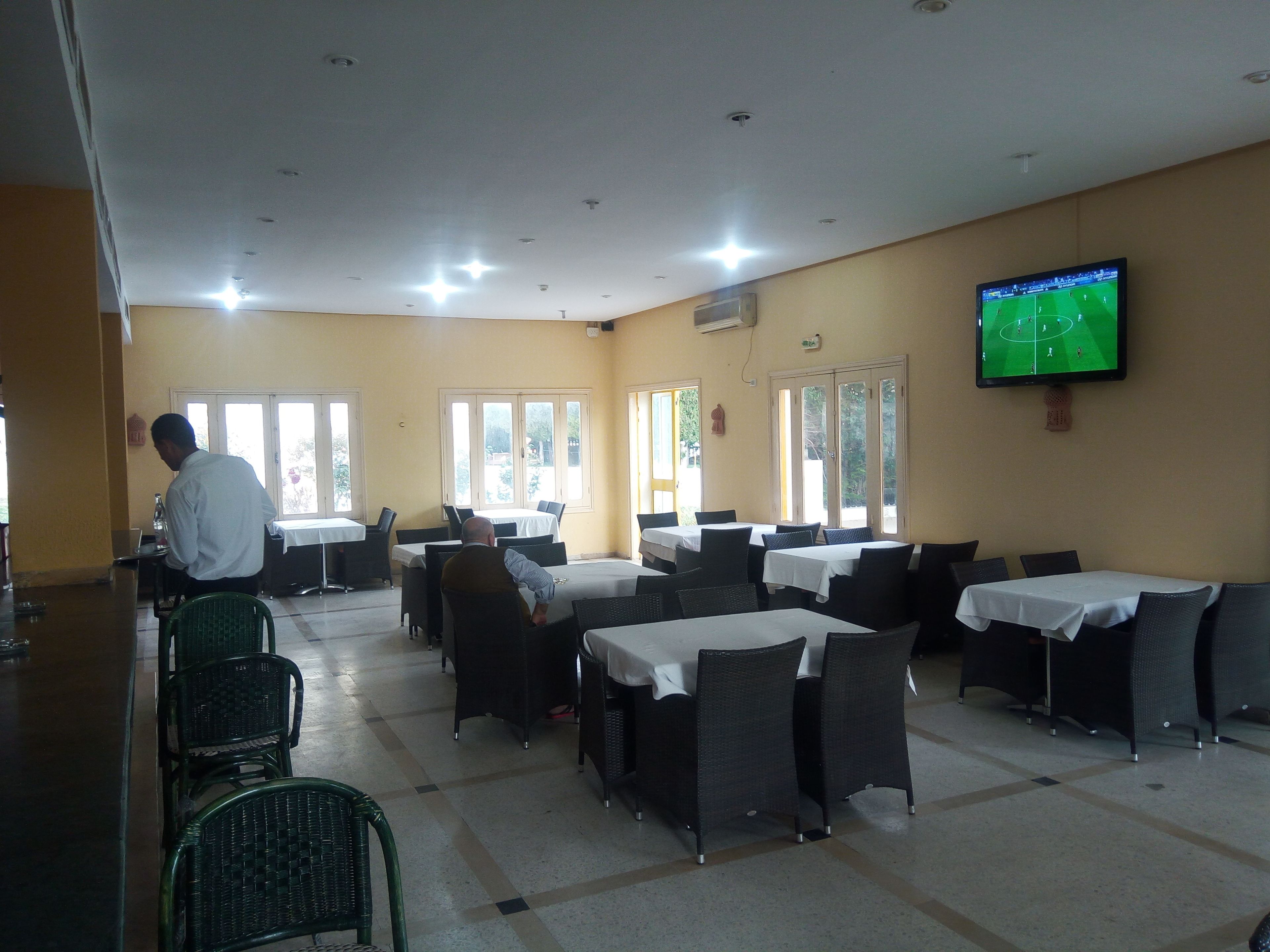 Un restaurante con mesas y sillas, una pantalla mostrando un partido de fútbol y un hombre atendiendo en la barra.