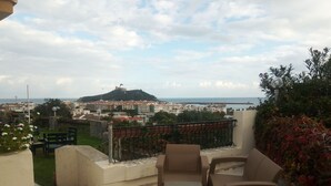 Una terraza con muebles de mimbre, vistas a un pueblo costero y una colina con una torre.