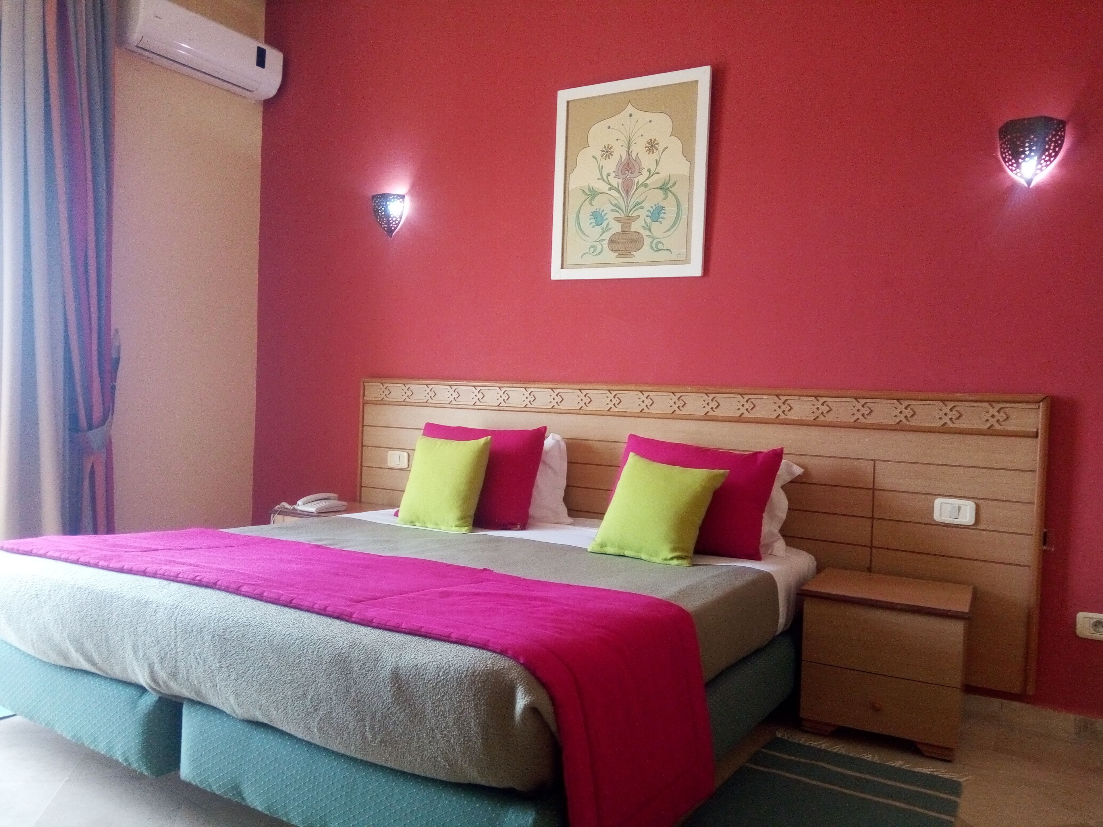Una habitación de hotel con cama, mesita de noche y aire acondicionado de pared. Se observa una obra de arte enmarcada sobre la cama.
