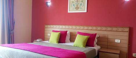 Una habitación de hotel con cama, mesita de noche y aire acondicionado de pared. Se observa una obra de arte enmarcada sobre la cama.