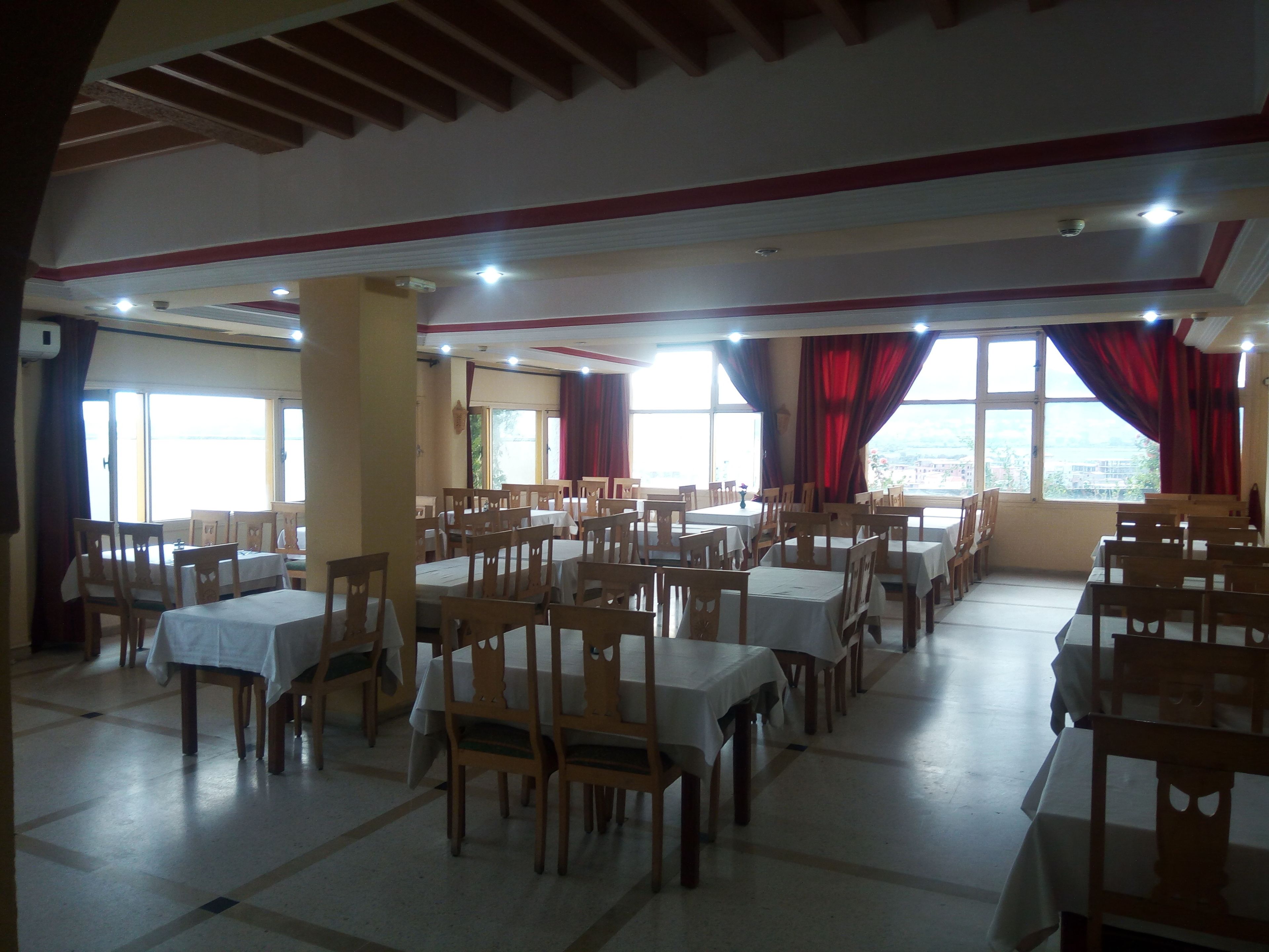Un'ampia sala da pranzo con diversi tavoli e sedie, grandi finestre con tende rosse e un soffitto alto con travi in legno.