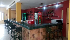 Una zona de bar con mostrador, taburetes, estantes con botellas y un refrigerador.