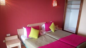 Una cama doble con una colcha color beige y rosa, dos mesitas de noche, una pequeña mesa, un tapete azul a rayas y un espejo en la pared.