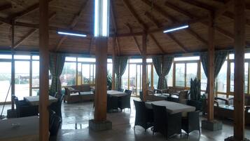 Amplio espacio interior con techos altos y vigas de madera, con varias mesas y sillas dispuestas para comer o socializar. Amplias ventanas permiten la entrada de luz natural y ofrecen vistas al exterior.