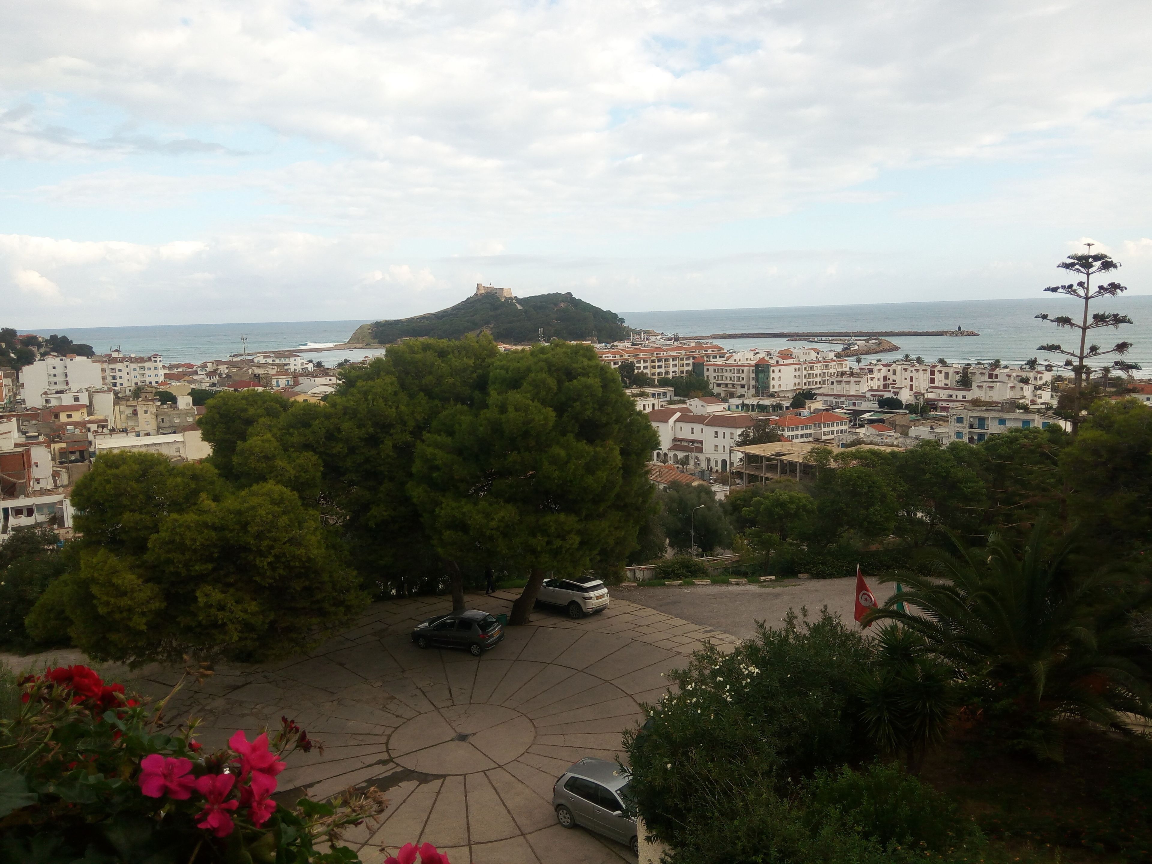 A coastal cityscape with a prominent hilltop fortress, surrounded by residential buildings and a clear view of the sea.