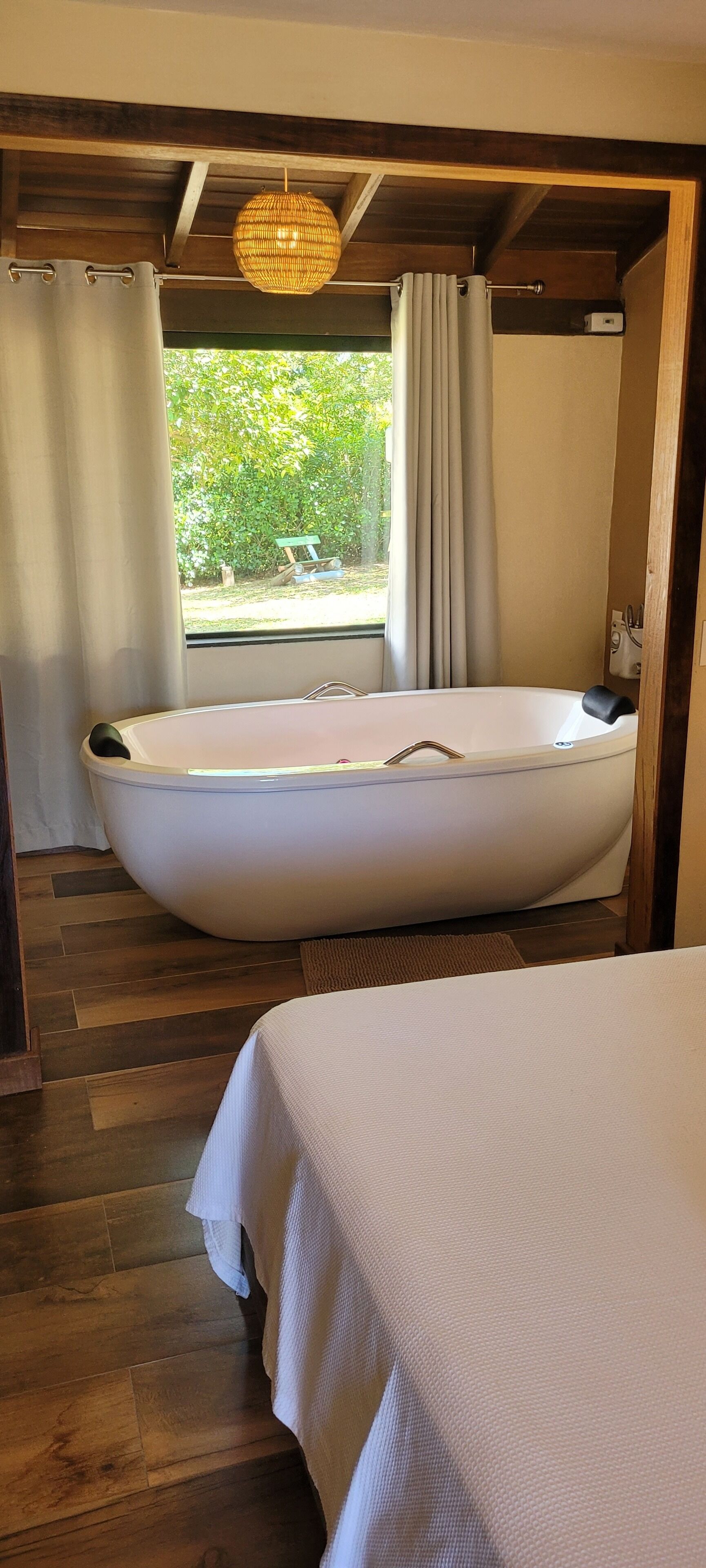 Una habitación con una bañera grande, piso de madera, una cama y un ventanal con cortinas.