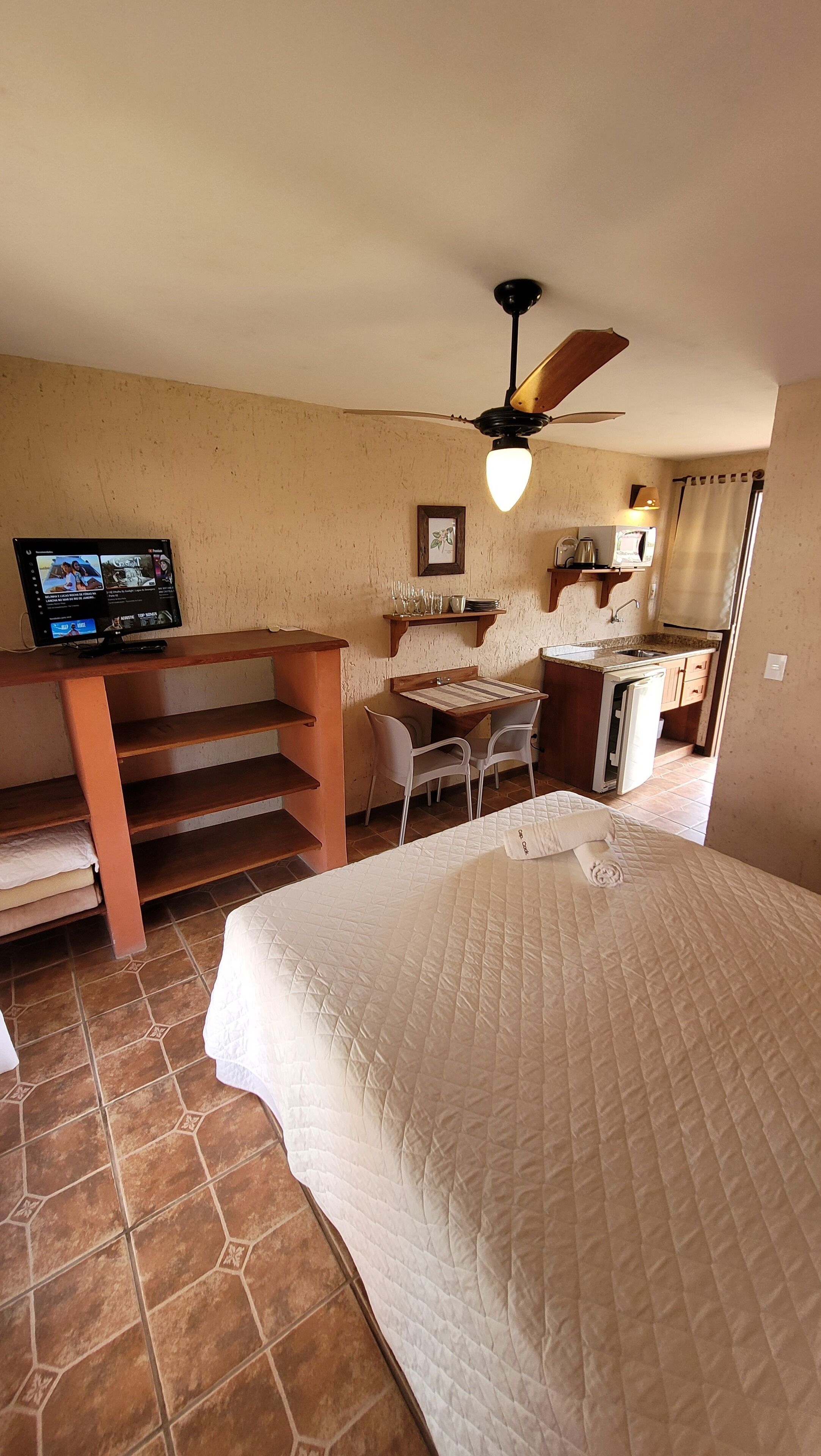 A compact room with a bed, a desk with a computer, a chair, a small kitchenette, and a ceiling fan.