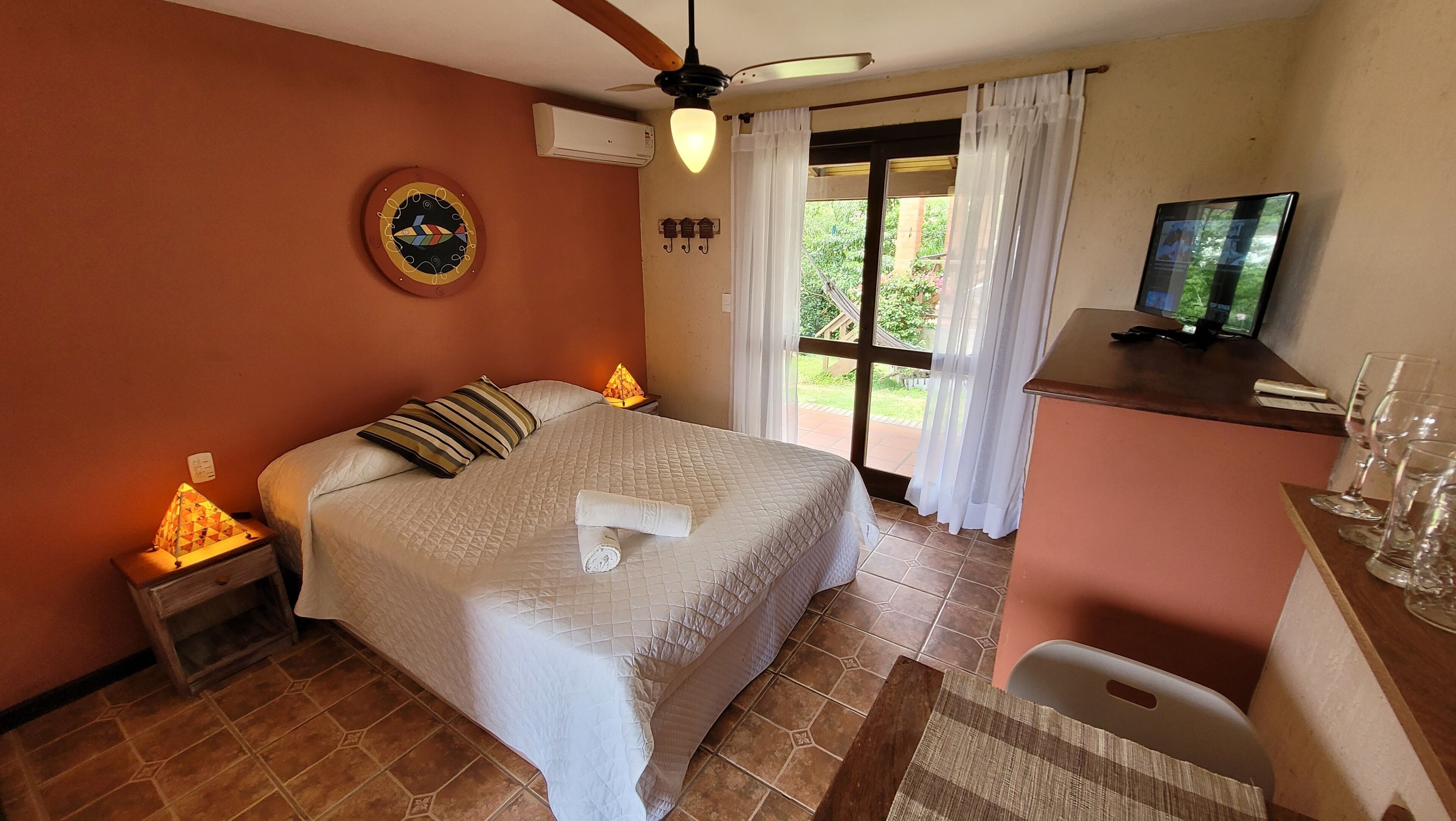 Quarto com cama, escrivaninha, televisão e janela com cortinas.