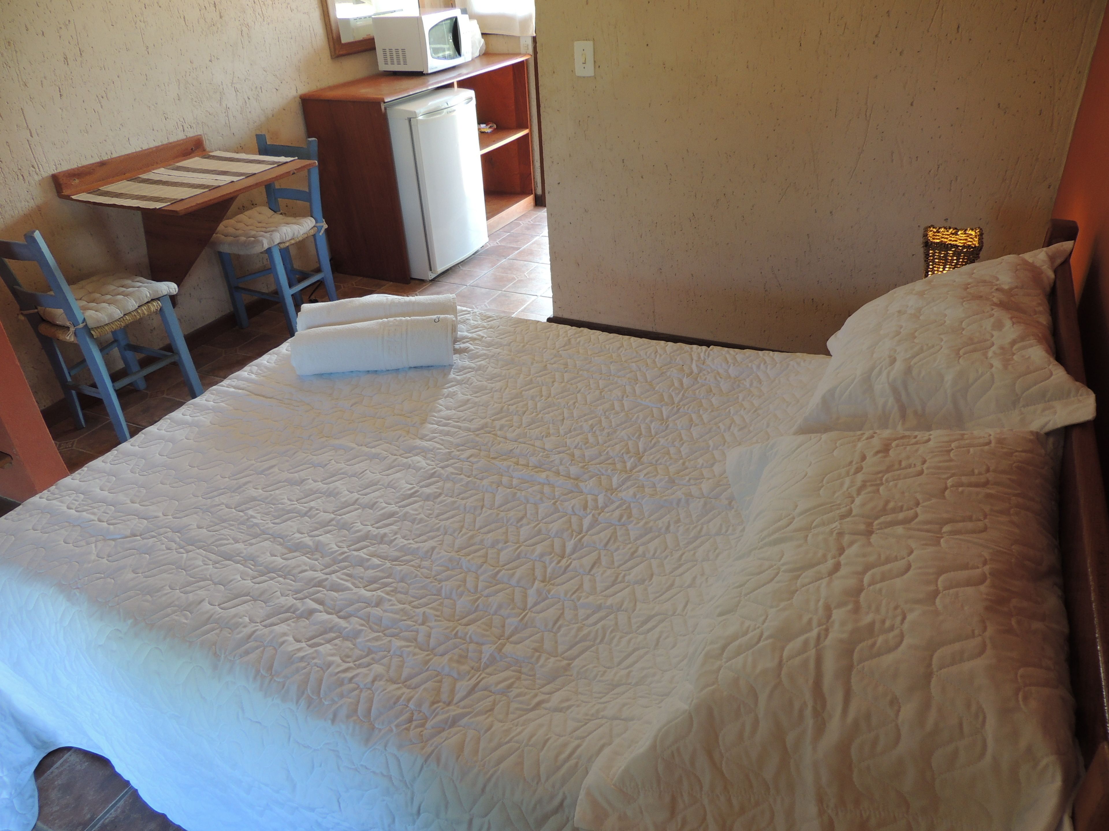 A bedroom with a large bed, a desk with a microwave, a chair, and a small refrigerator.
