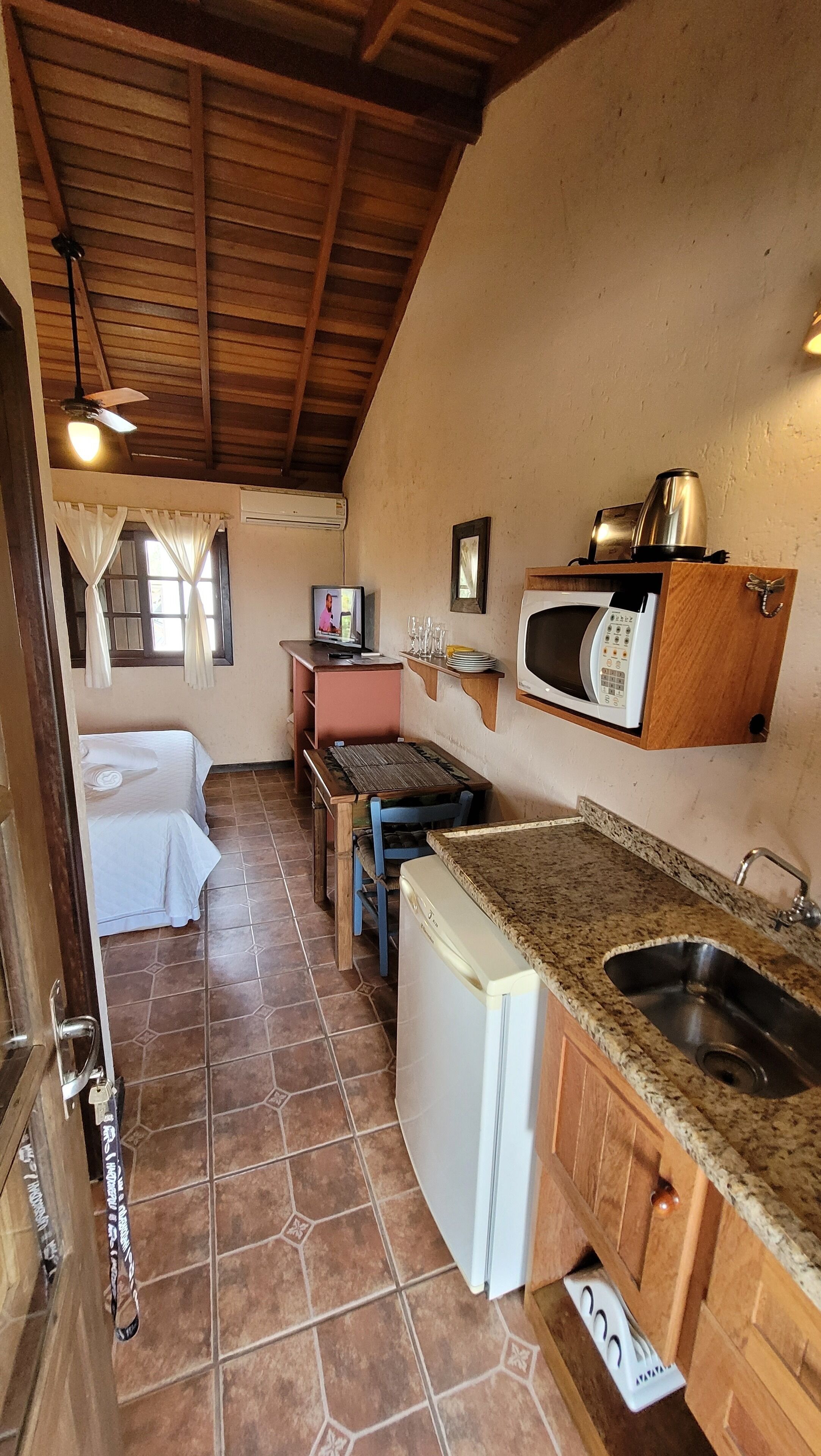 Cocina compacta con microondas, pileta y heladera pequeña. Tiene techo de madera y ventana con cortinas.