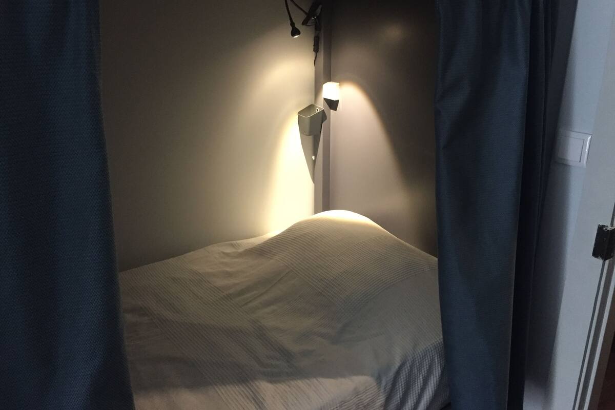 A bed with a white mattress and a single pillow, a bedside lamp, and a curtain partially drawn.