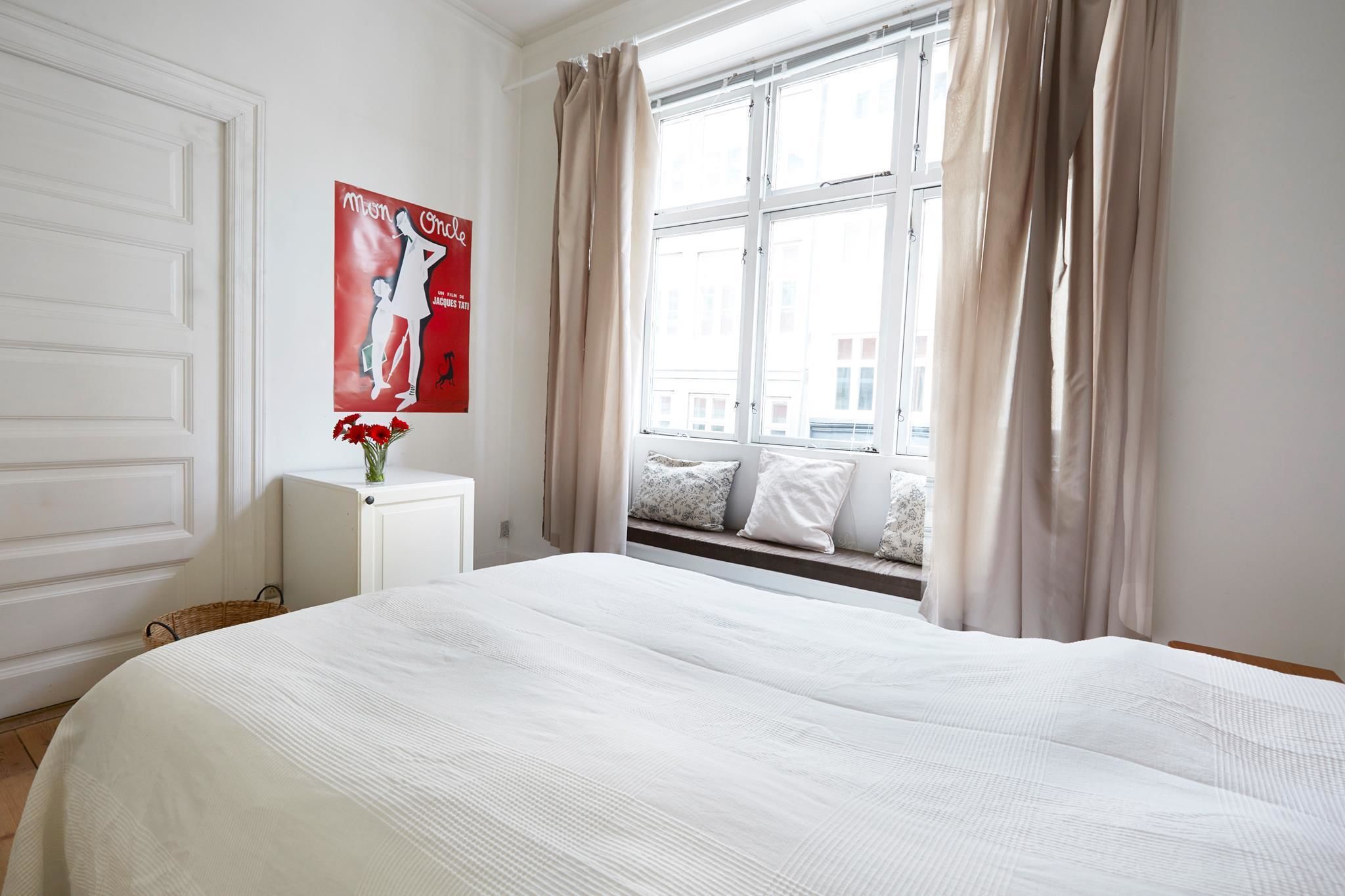 A bedroom with a large bed, a window with curtains, a radiator, a poster on the wall, and a small table with a vase of flowers.