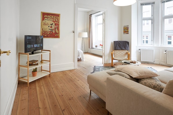 A living room with a wooden floor, a beige sofa, a TV on a wooden shelf, and a poster on the wall.