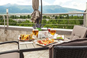 Un balcon avec une table dressée pour un repas, où l’on aperçoit des assiettes de nourriture et des verres de bière, donnant sur un paysage montagneux.