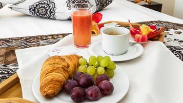 Un desayuno con croissant, uvas, un vaso de jugo de naranja y una taza de café sobre una cama con almohadillas estampadas.