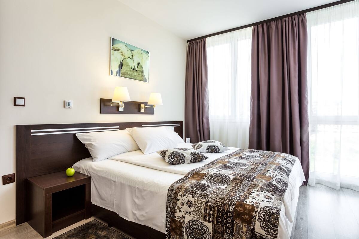 A neatly made bed with a patterned bedspread, a bedside table with a lamp, a wall-mounted light fixture, and a painting above the bed.