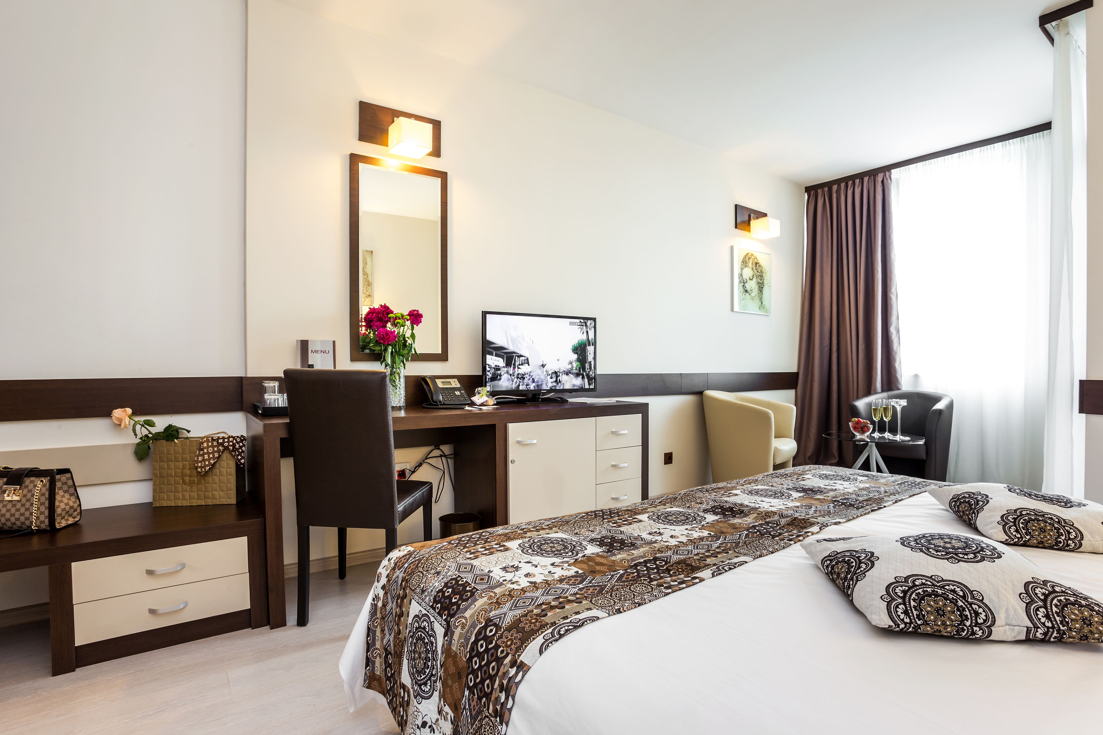 Una habitación de hotel con cama, escritorio, silla, computadora y ventana con cortinas.