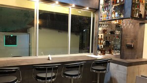 A bar area with a long counter, three bar stools, and a shelf with various bottles and glasses. - Kay's Riverview Resort (Arpora)