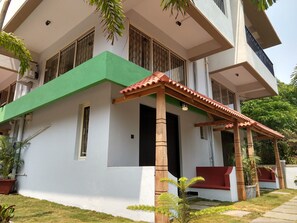 Um edifício de dois andares com fachada verde e branca, varanda coberta com telhado vermelho e um pequeno jardim com plantas em vasos.