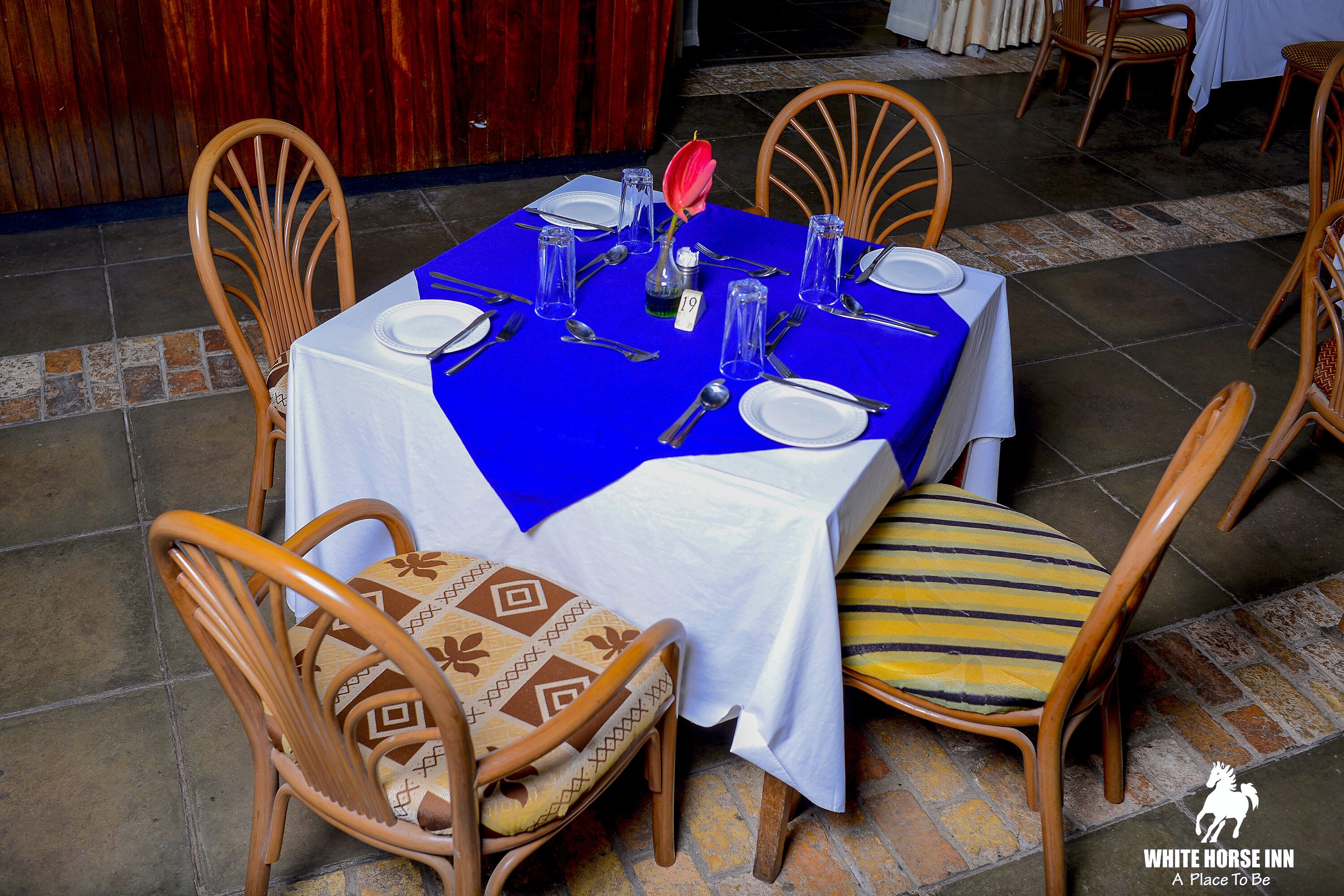 Une table à manger dressée avec une nappe bleue, des chemins de table blancs et des chaises en bois ornées de coussins à motifs.