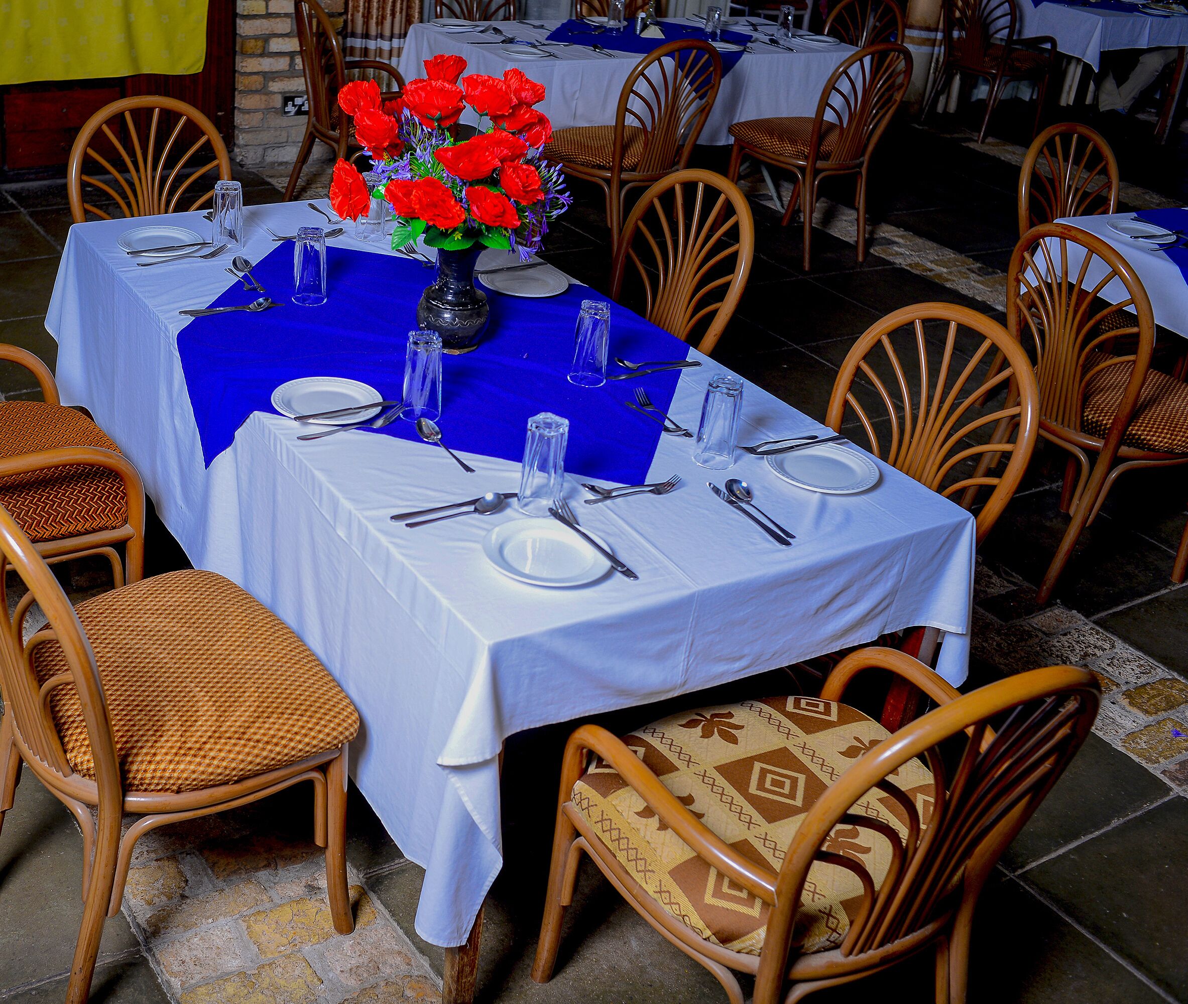 A dining table set with white tablecloths, blue table runners, and a vase of red flowers.