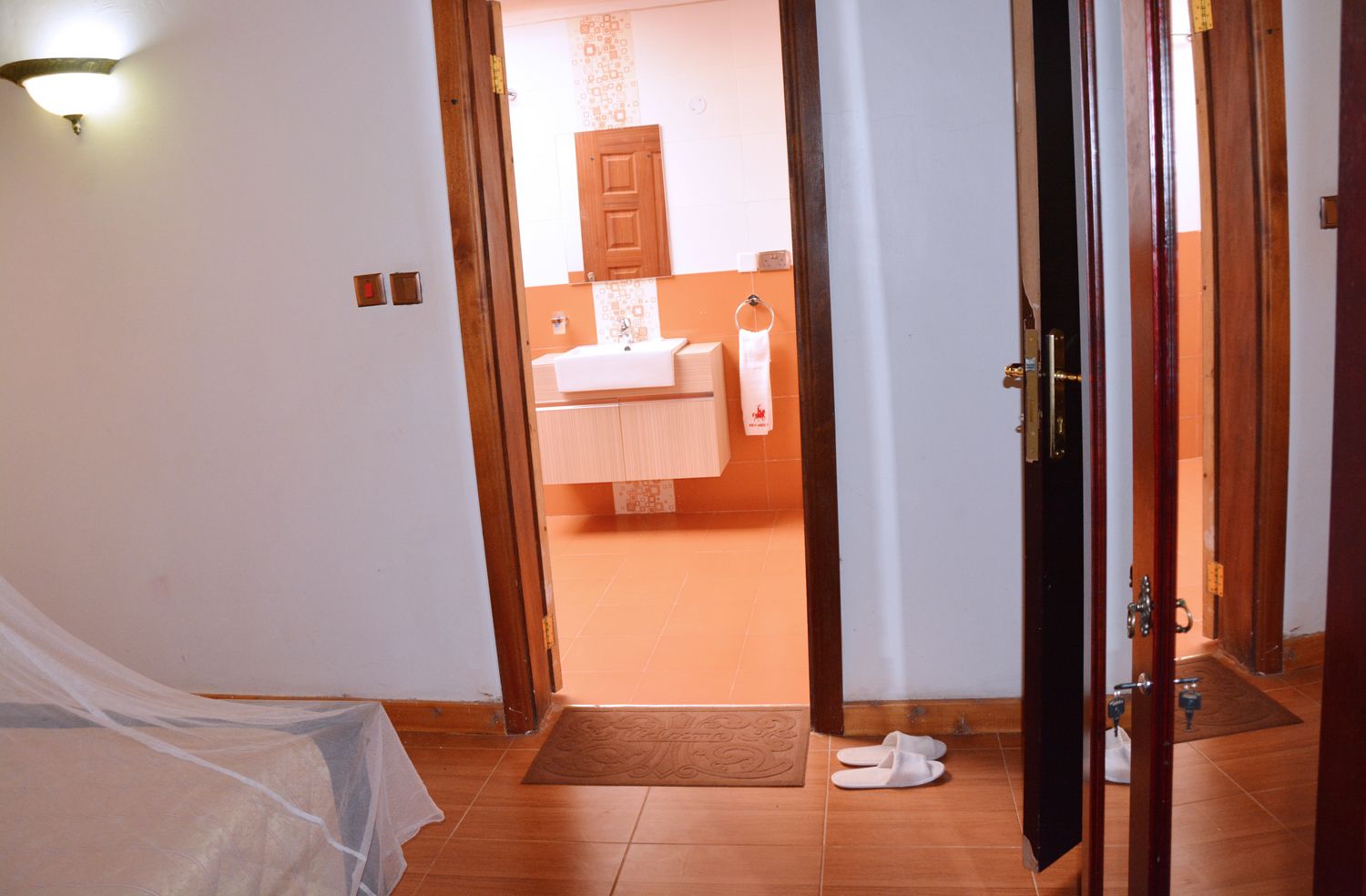 A room with a bed, a bedside lamp, and a door leading to a bathroom with a sink and towel rack.