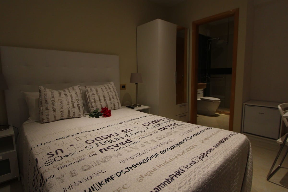 Um quarto com uma cama, uma mesa de cabeceira, uma luminária, um guarda-roupa e uma escrivaninha com uma cadeira.