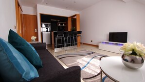 Una sala moderna con un sofá, una mesa de centro con un jarrón de flores y una zona de cocina con televisor y taburetes de bar.
