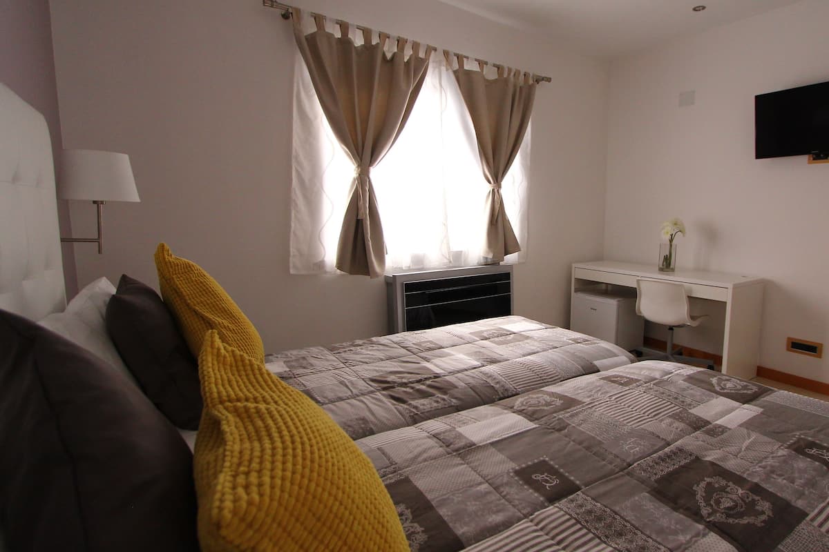 Quarto com cama, escrivaninha, televisão e janela com cortinas.