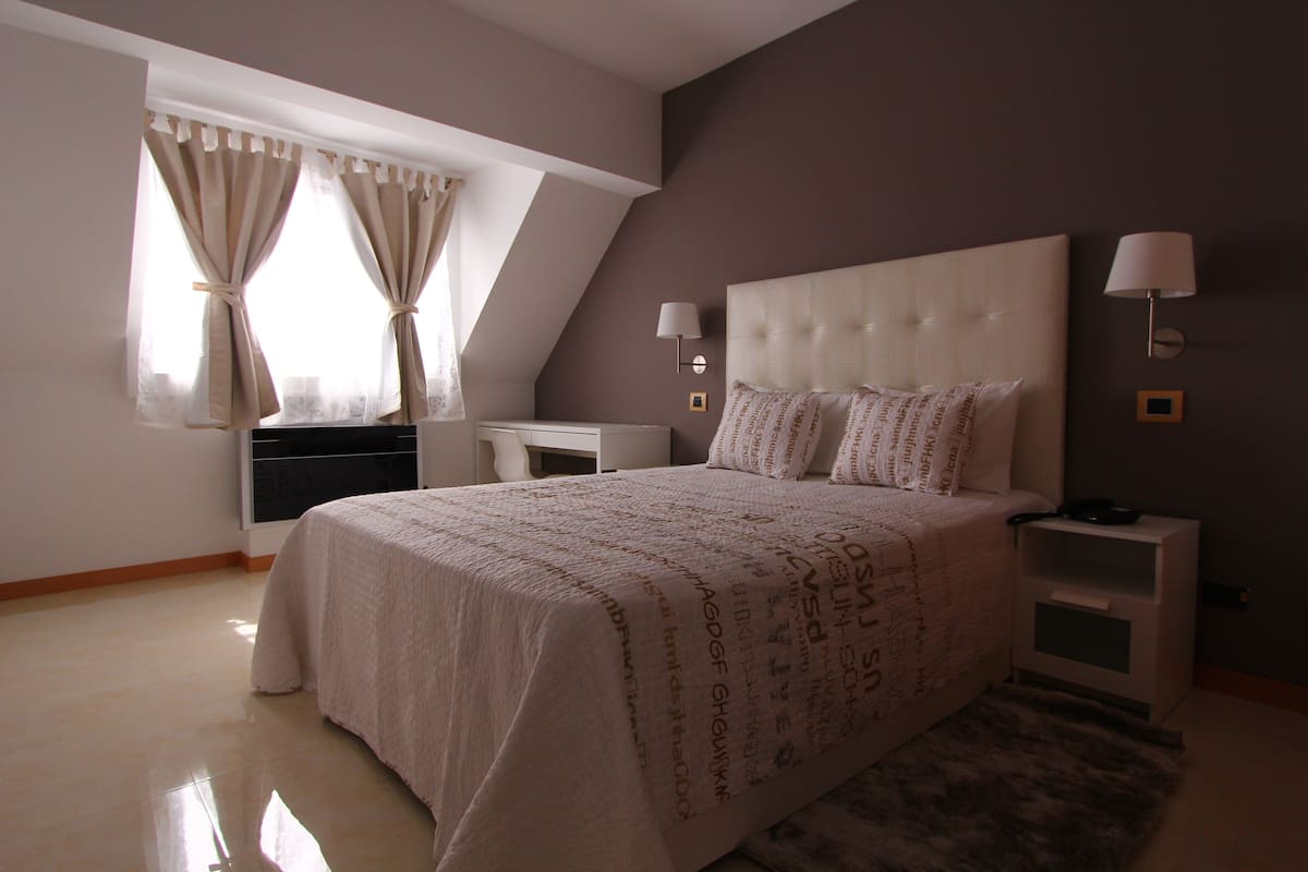 Quarto com cama grande, duas mesas de cabeceira, um criado-mudo, janela com cortinas e luminária na parede.