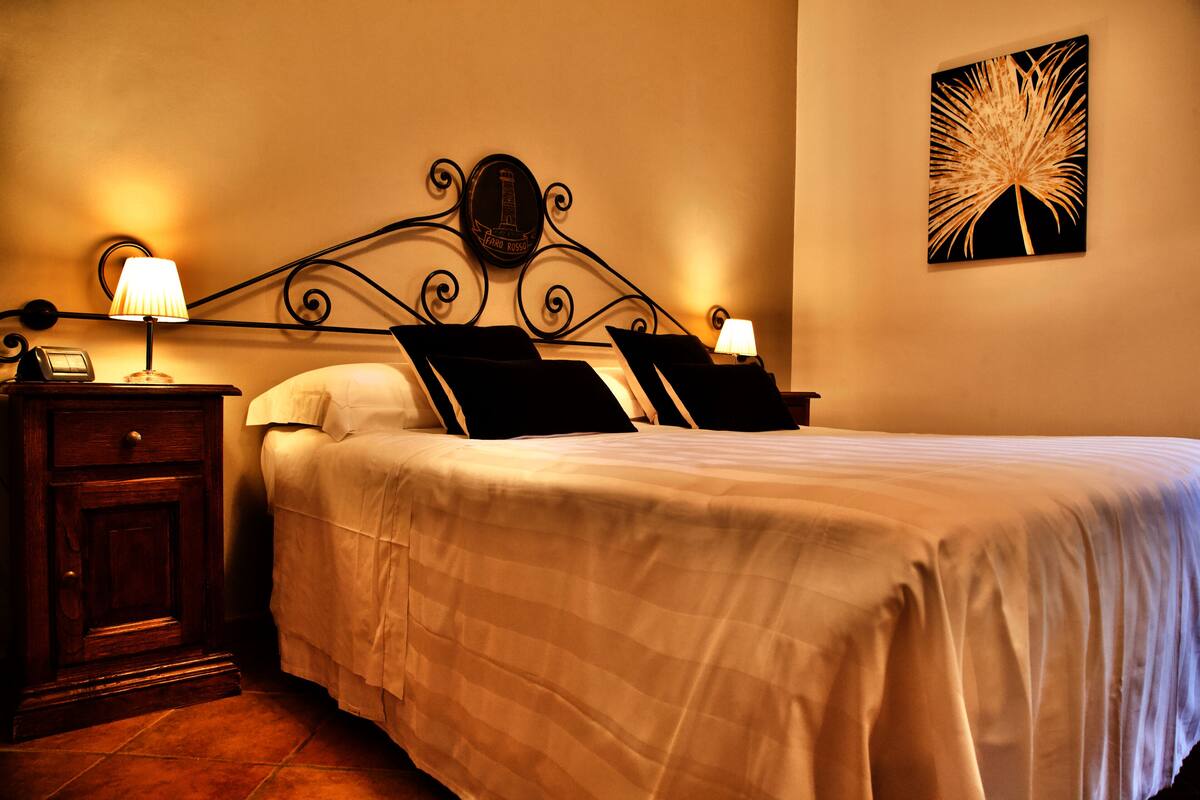 A neatly made bed with white linens and black pillows, a bedside table with a lamp, and a wall-mounted artwork.