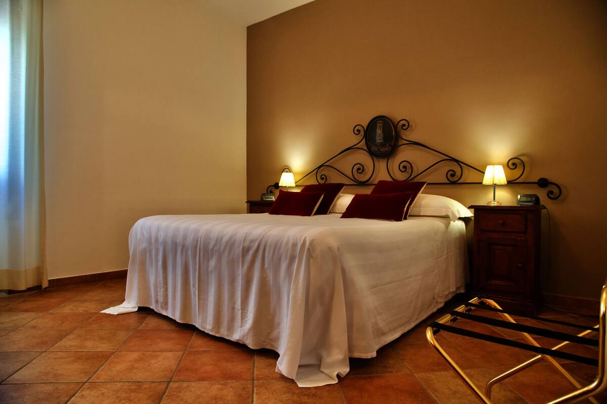 A neatly made bed with white linens and red pillows, a wooden nightstand with a lamp, and a metal chair.