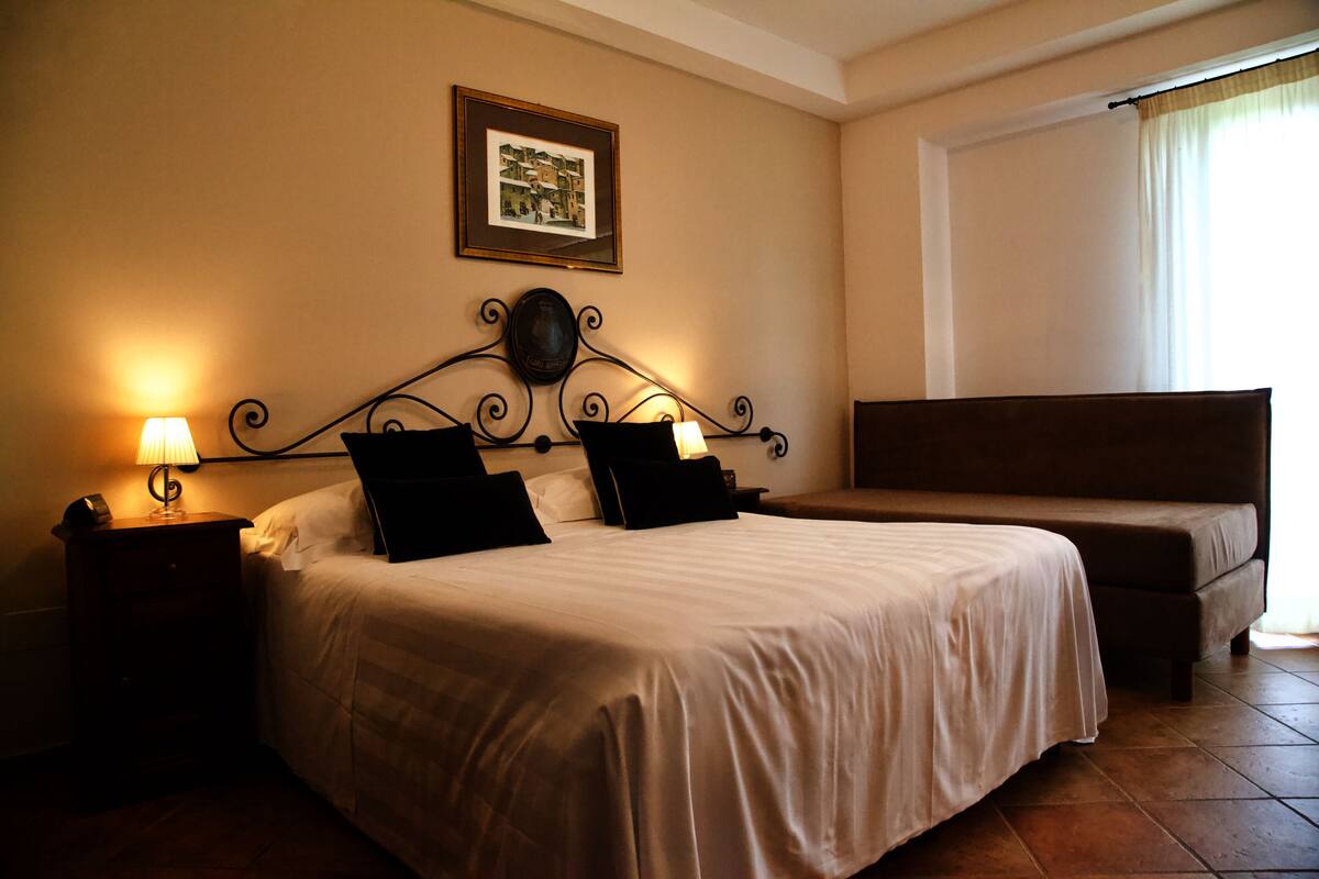 A hotel room with a large bed, a bedside table with a lamp, a framed picture on the wall, and a window with curtains.