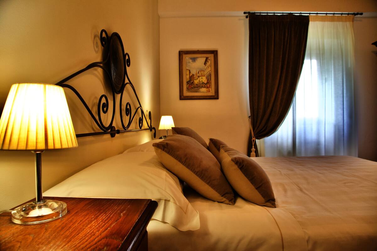 A hotel room with a bed, two bedside lamps, a painting on the wall, and a window with curtains.