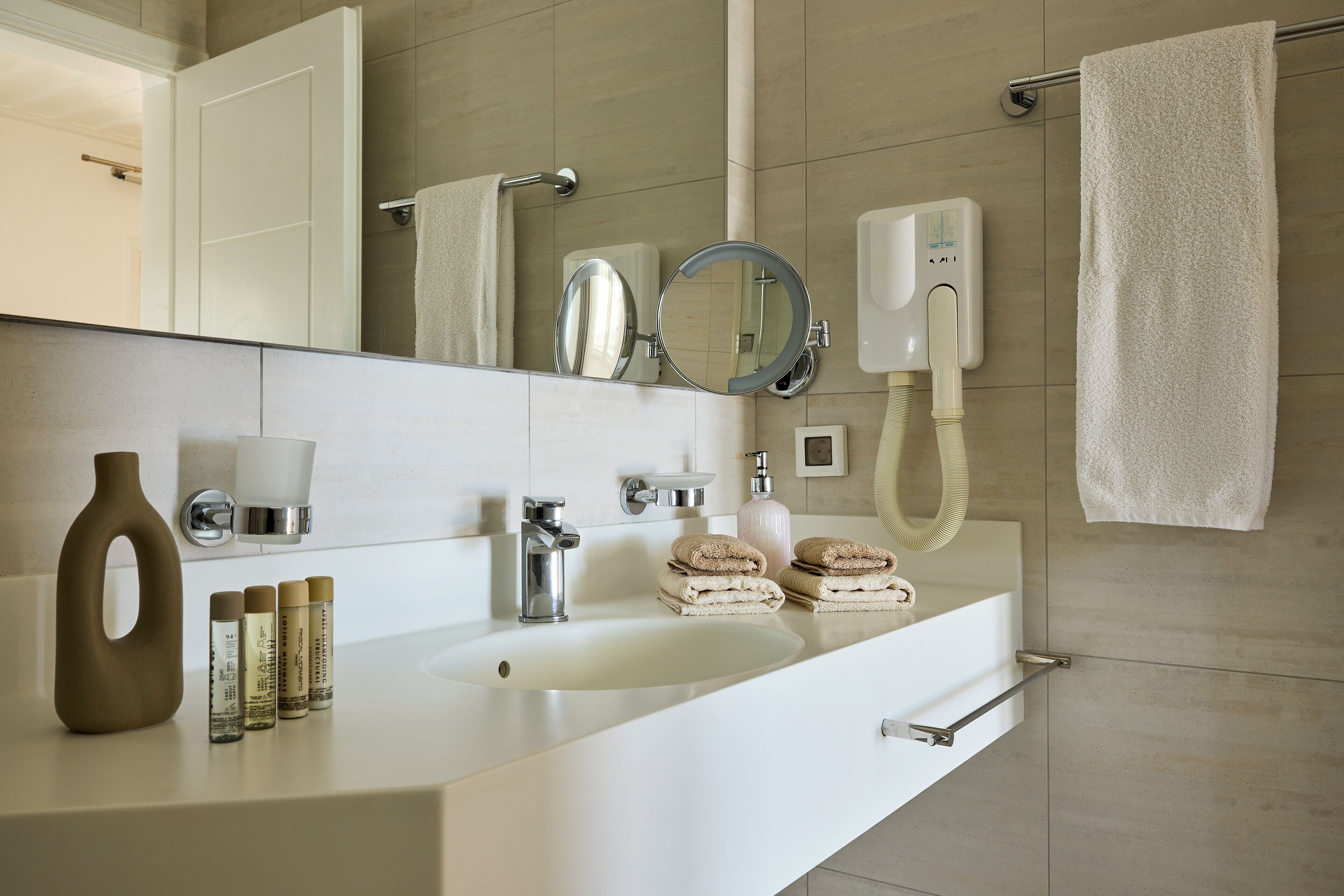 A modern bathroom with a white sink, mirror, and towel rack. There are toiletries on the counter and a showerhead above the sink.