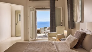 A bedroom with a bed, a chair, a lamp, and a view of the sea through an open door. - Selana Suites (Sifnos)