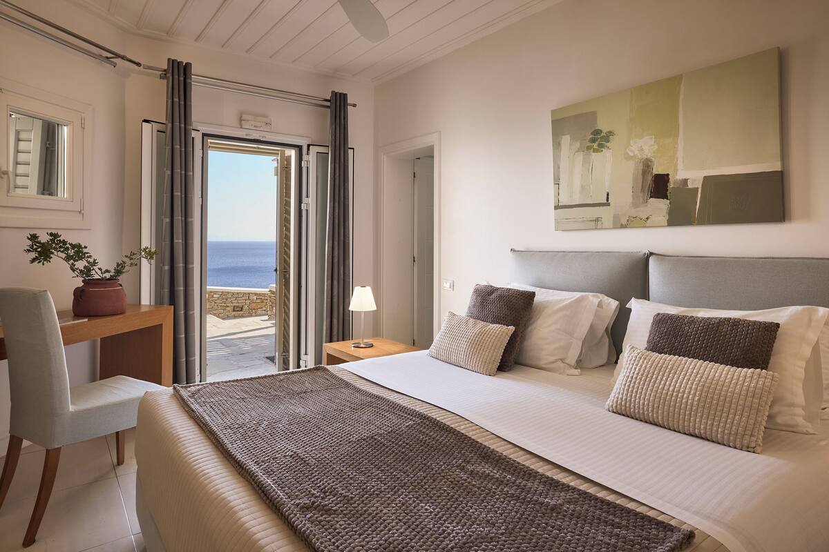 A bedroom with a bed, a desk, a chair, a nightstand, and a view of the sea.
