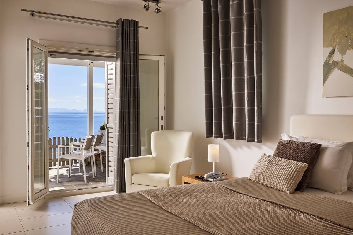 A bedroom with a bed, a chair, a nightstand, and a view of the sea through a sliding door.